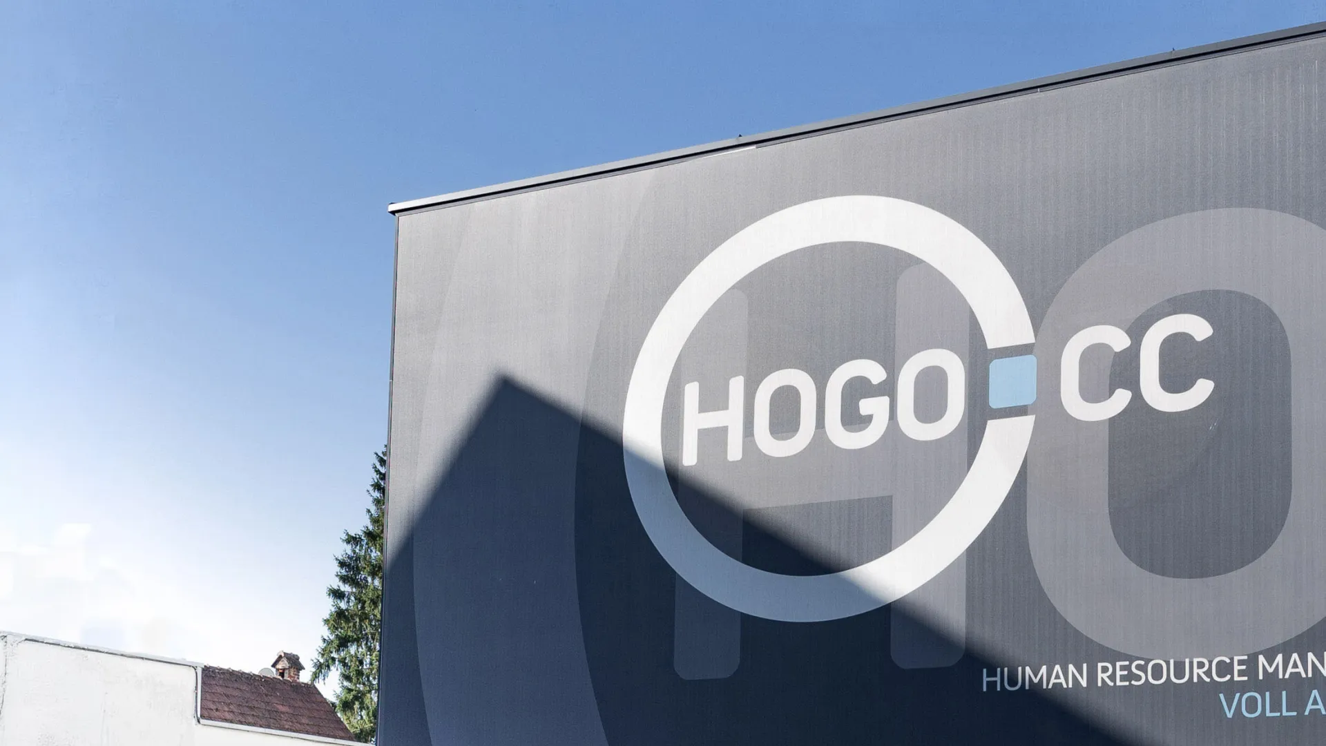 Werbeplakat mit großem HOGOCC-Logo und Teilansicht eines Rennwagens vor blauem Himmel.